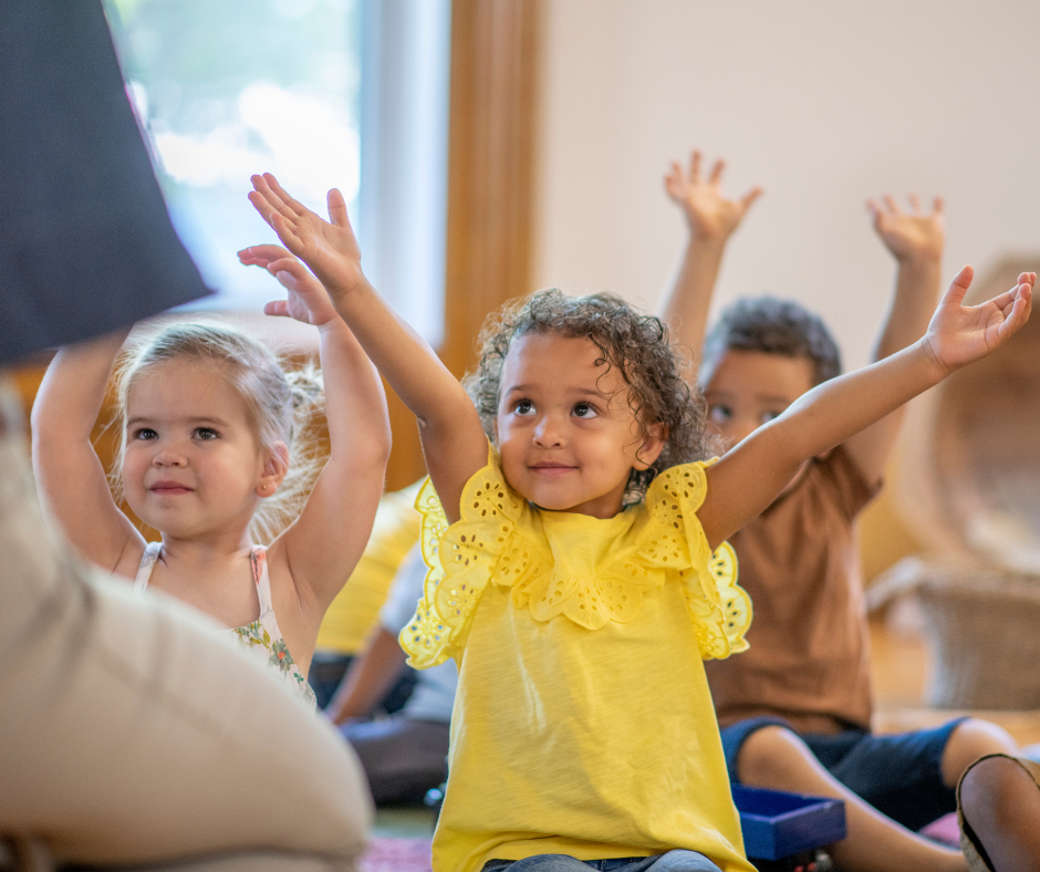 Children in a classroom stretching their arms over their heads and smiling at the teacher. 