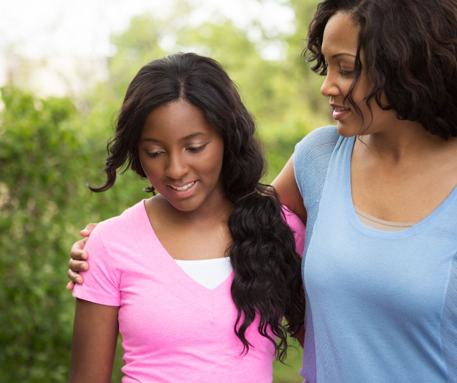 Two women stand together in a park, discussing summer camp prep ideas.