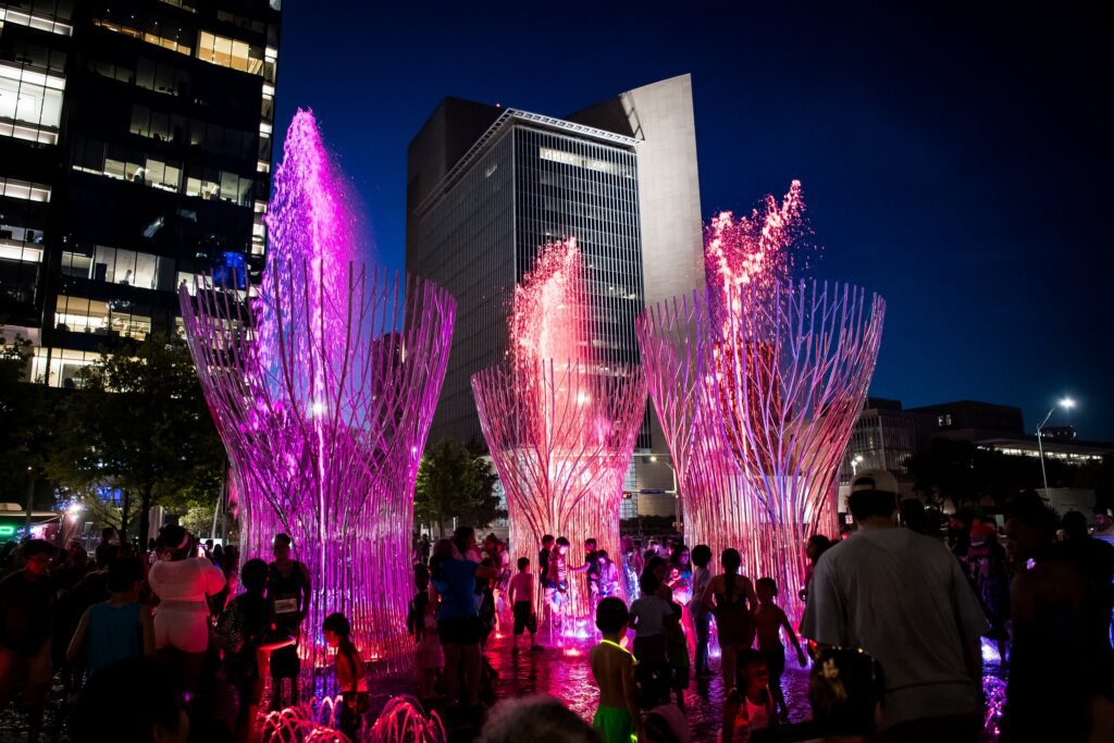 Families and friends stand around a colorful fountain at Klyde Warren Park, creating a lively atmosphere at night in Dallas.